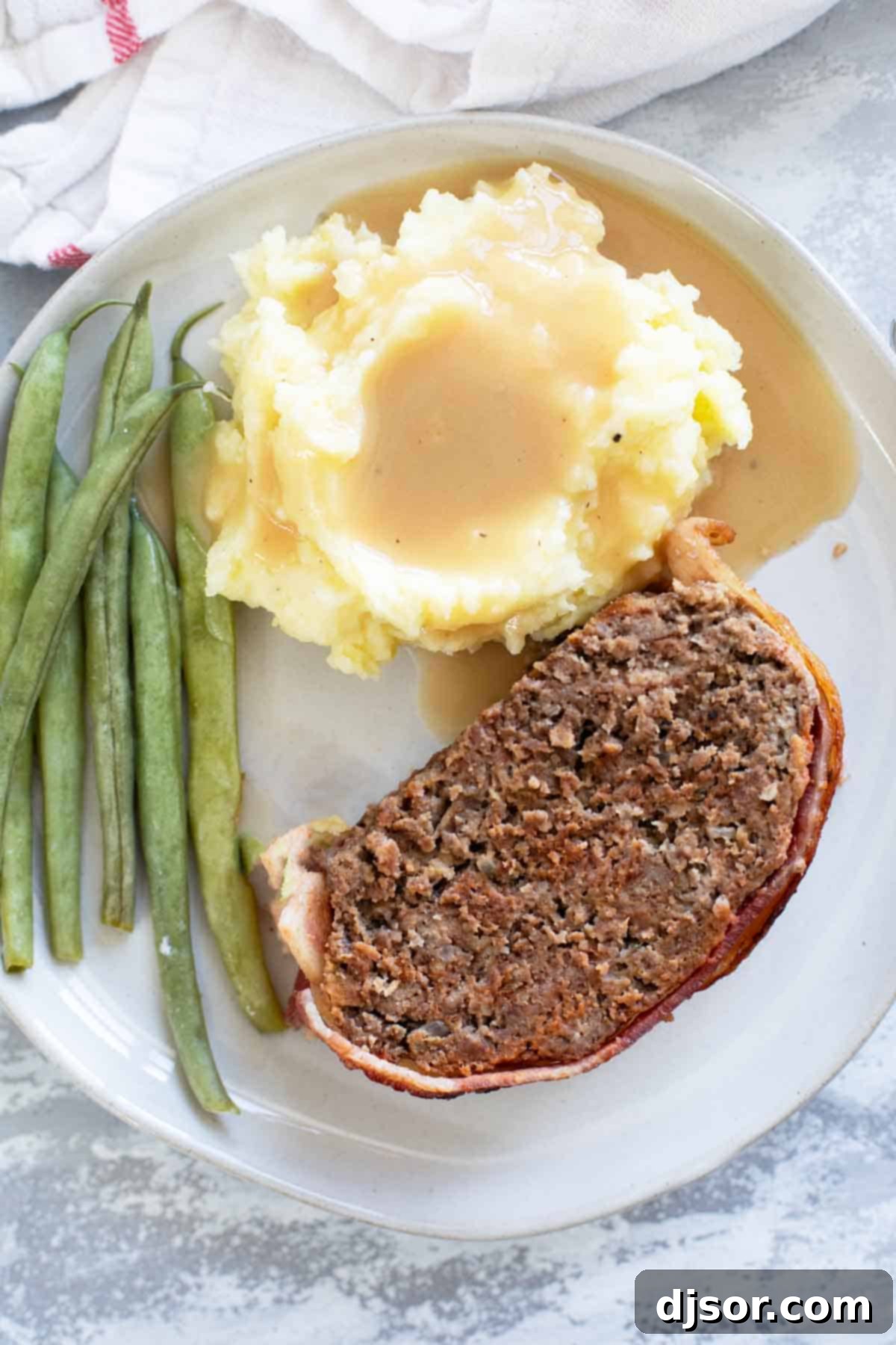 The Ultimate Bacon Meatloaf 8 Plate with a slice of bacon wrapped meatloaf, mashed potatoes and gravy, and green beans.
