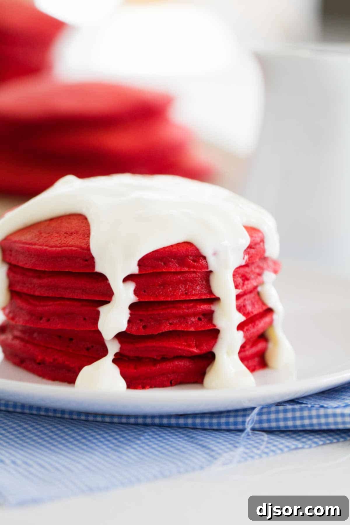 Red Velvet Pancakes with Cream Cheese Icing A generous stack of Red Velvet Pancakes on a white plate, lavishly topped with a creamy cream cheese icing and a light dusting of powdered sugar. The vibrant red color of the pancakes contrasts beautifully with the white icing, making it an enticing breakfast or brunch option.