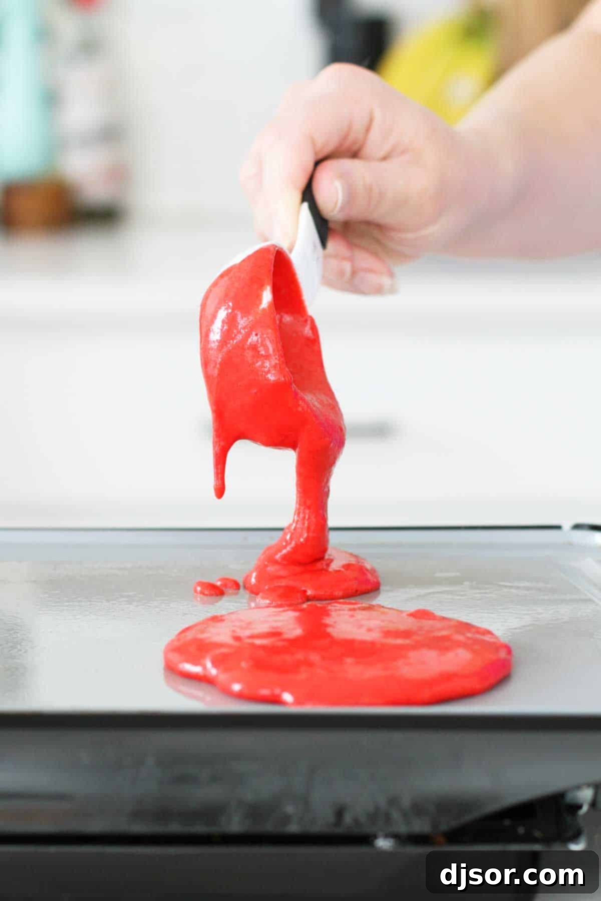 Pouring Red Velvet Pancake Batter A chef's hand carefully pouring vibrant red velvet pancake batter onto a hot griddle, illustrating a key step in the cooking process. The rich red color of the batter stands out against the dark surface of the griddle, promising delicious results.